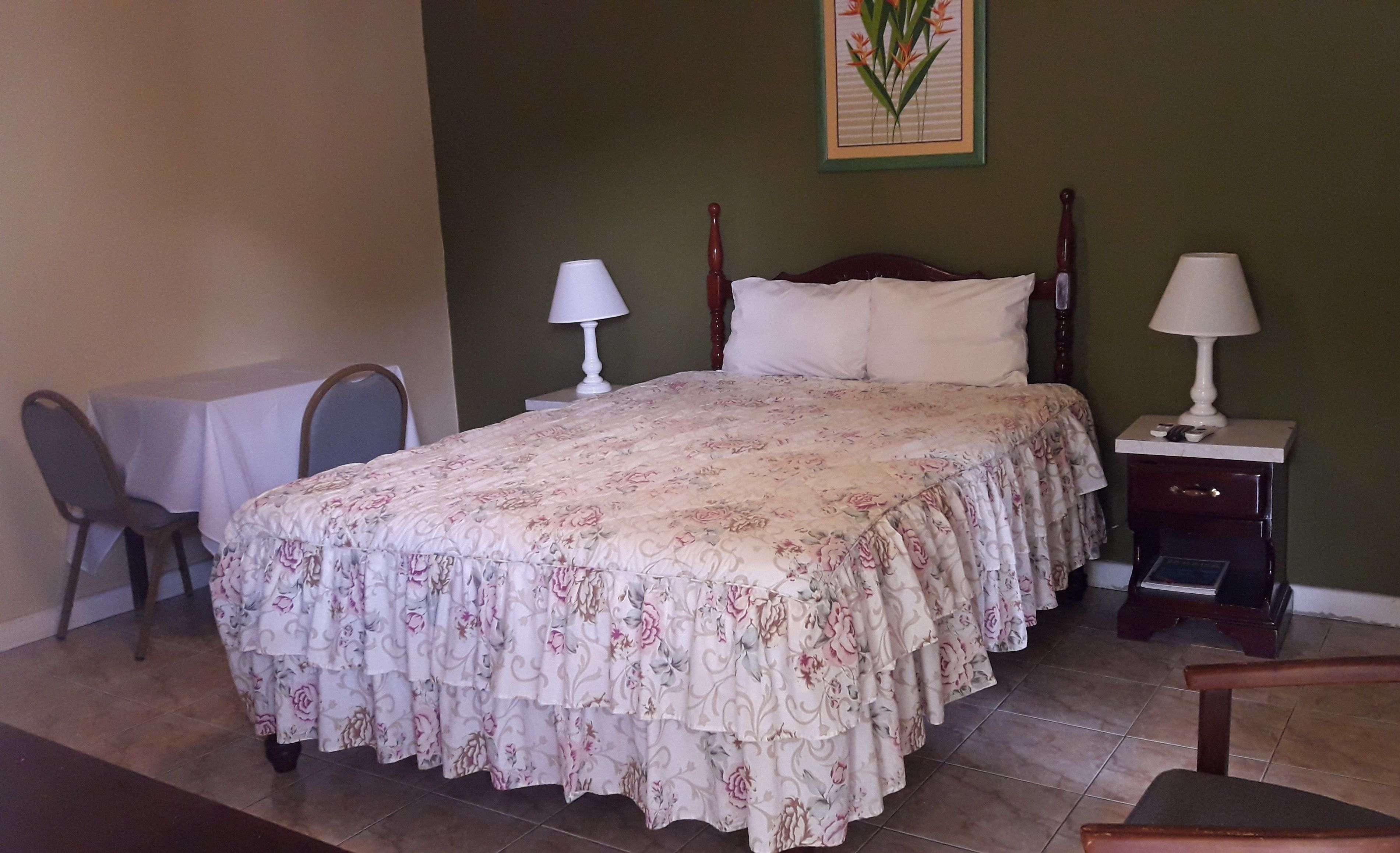 A neatly made bed with floral bedding, a nightstand with a lamp, a chair, and a table with a tablecloth.