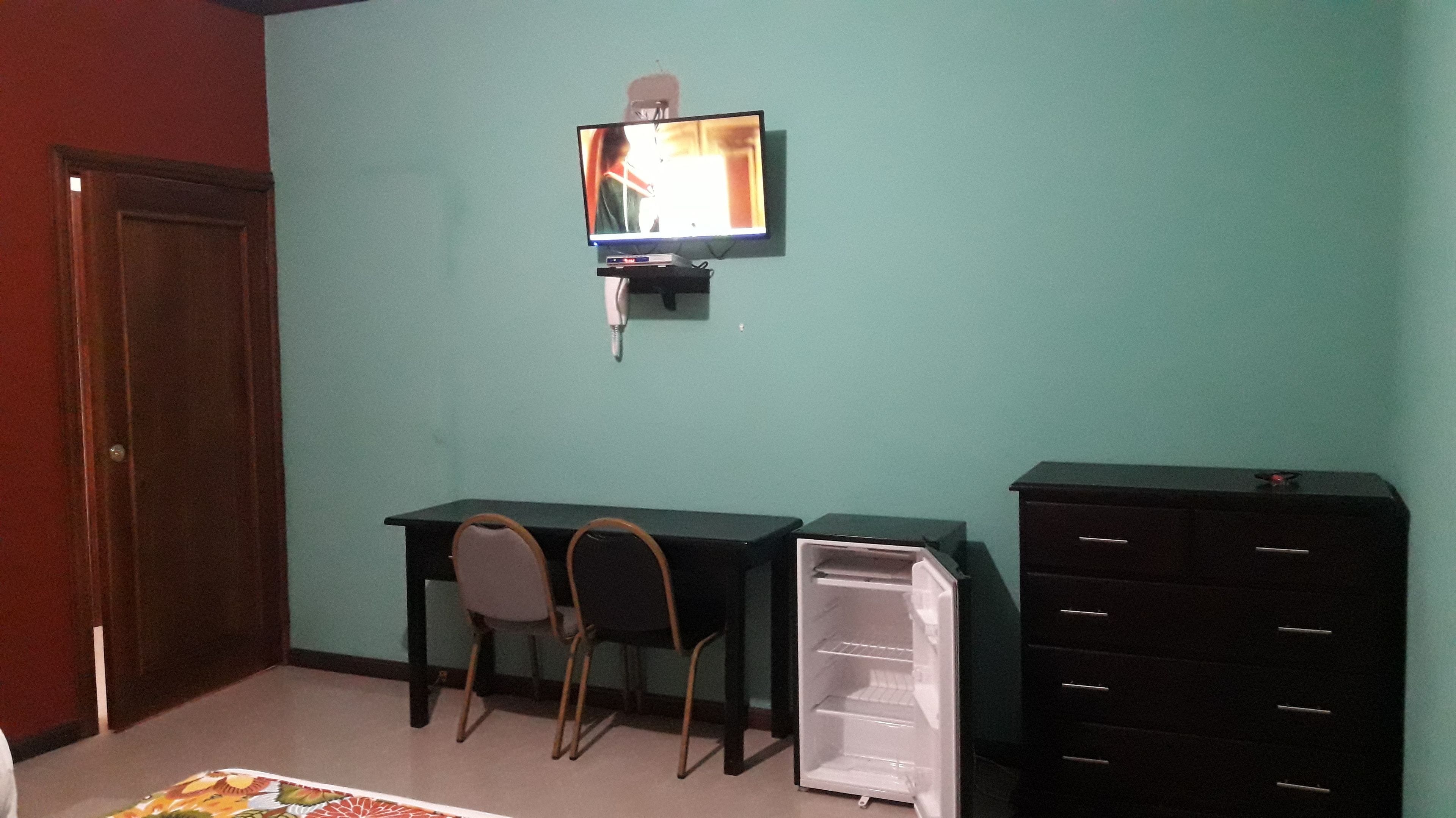 Una habitación con un escritorio, dos sillas, un televisor fijado a la pared y un aparador negro.