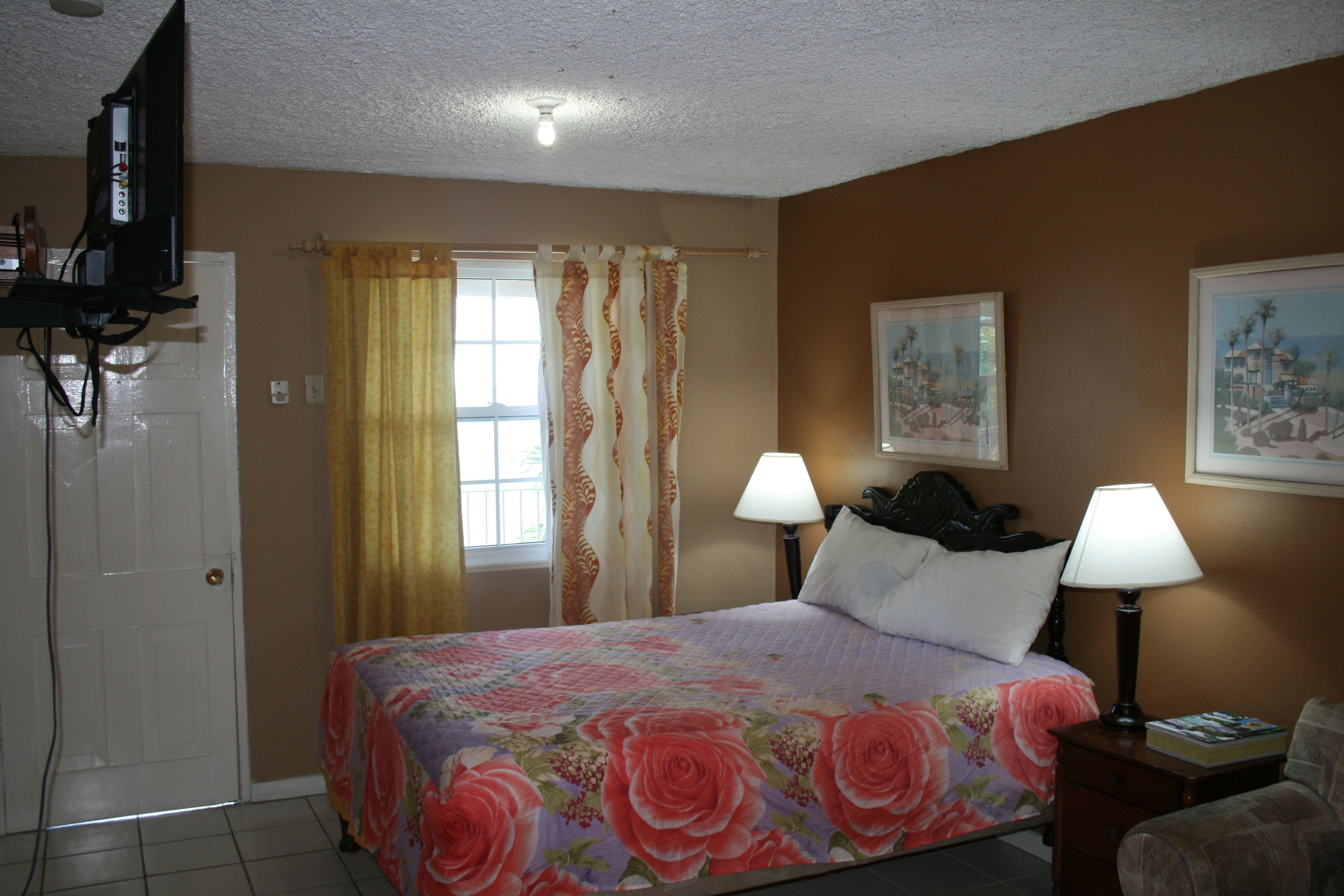A hotel room with a bed, two bedside lamps, a TV, a window with curtains, and a painting on the wall.