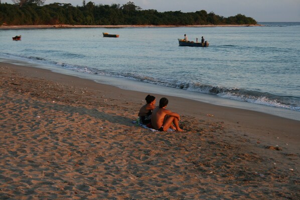 A beach with two people sitting on the sand, boats in the water, and a forested area in the background.