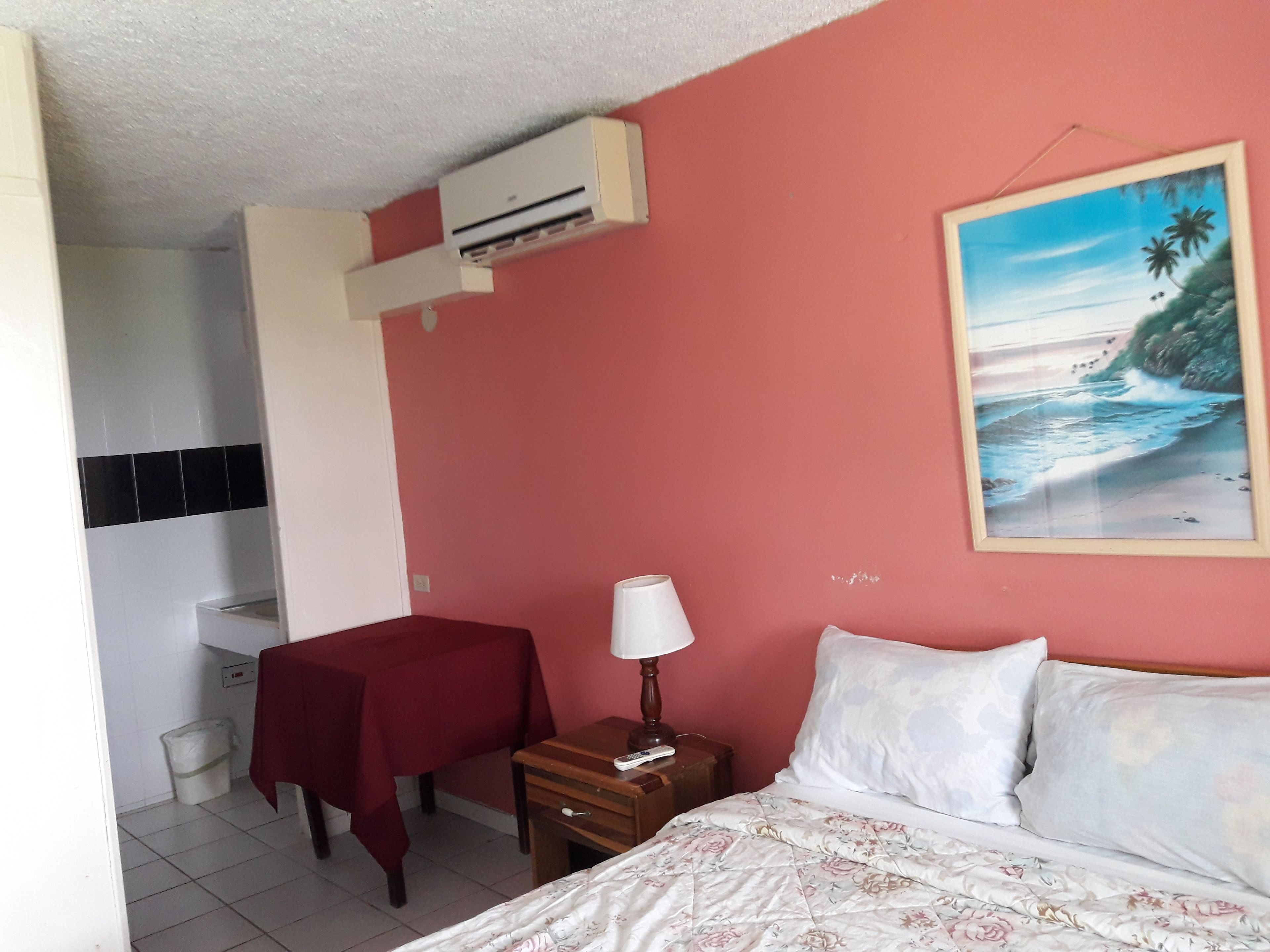 Una camera da letto con un letto, un comodino, un tavolo con tovaglia rossa, un condizionatore d'aria a parete e un quadro raffigurante una spiaggia.