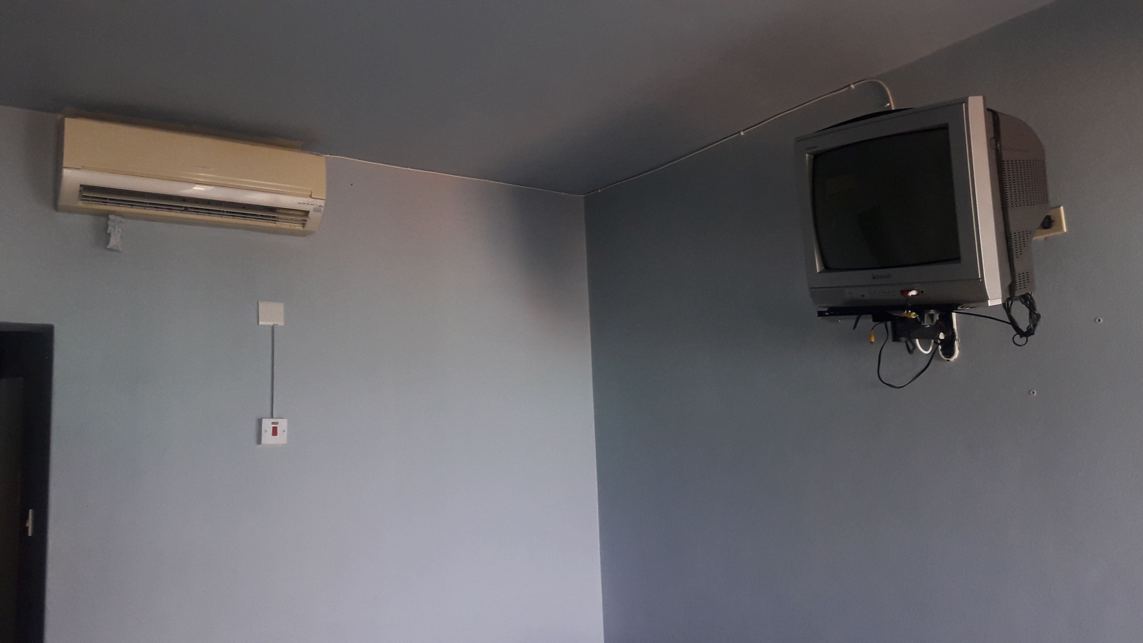 A room with a mounted television on the wall, an air conditioning unit, and a power outlet.