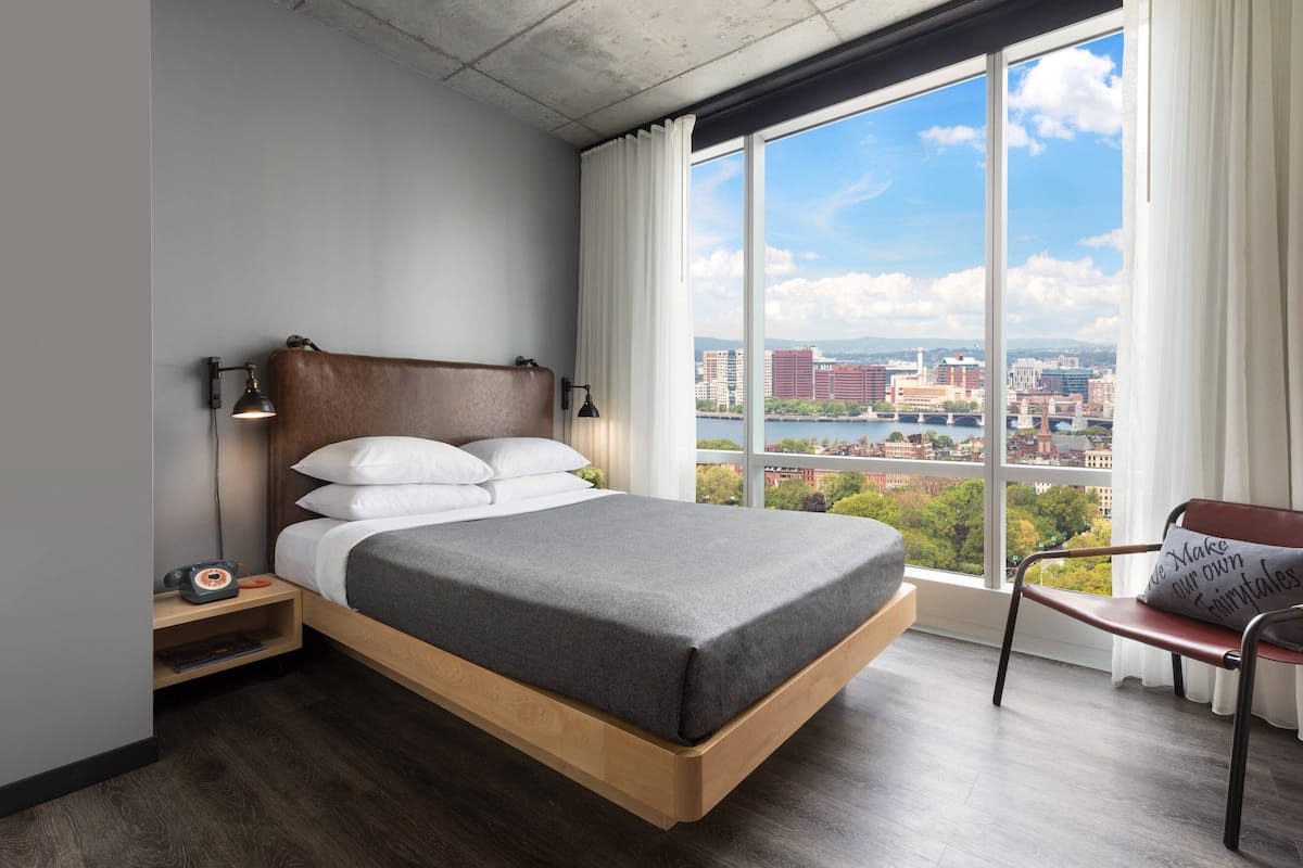 A modern hotel room with a large bed, a nightstand with a lamp, a chair, and a view of the city through large windows.