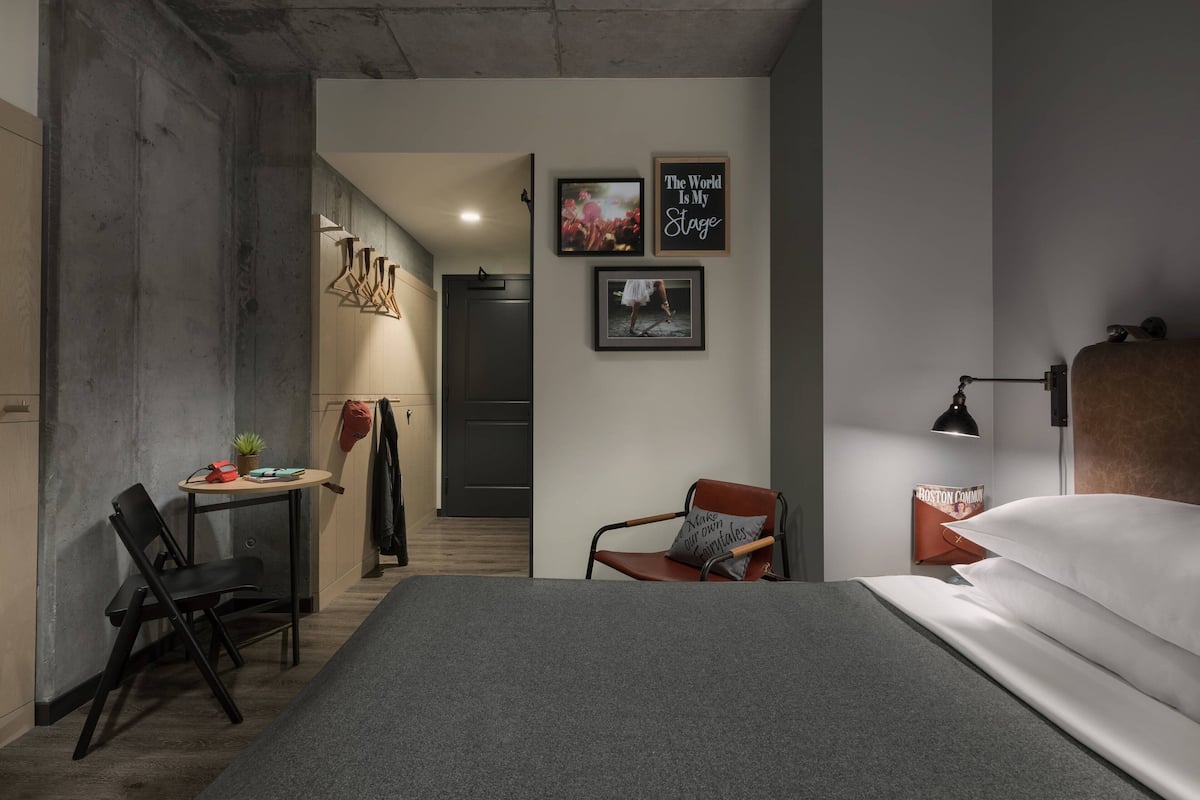 A modern hotel room with a bed, a desk with a chair, a small table with a plant, and a wall with framed pictures and a sign.