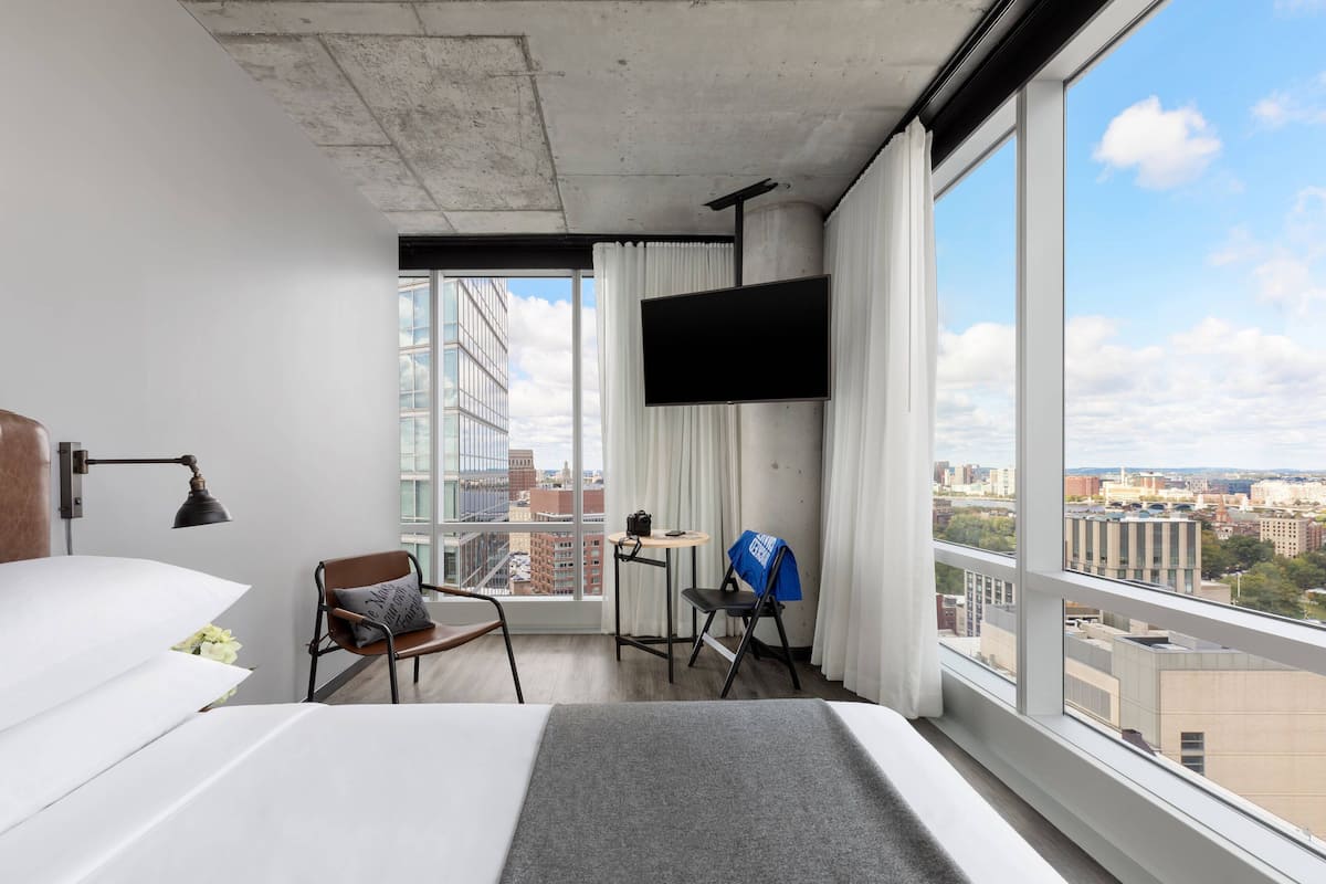 A hotel room with a large bed, a desk with a chair, a TV mounted on the wall, and a view of the city through large windows.