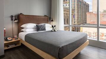 A modern hotel room with a large bed, a nightstand with a lamp, a small table with a telephone and a mug, and a view of the city through large windows.