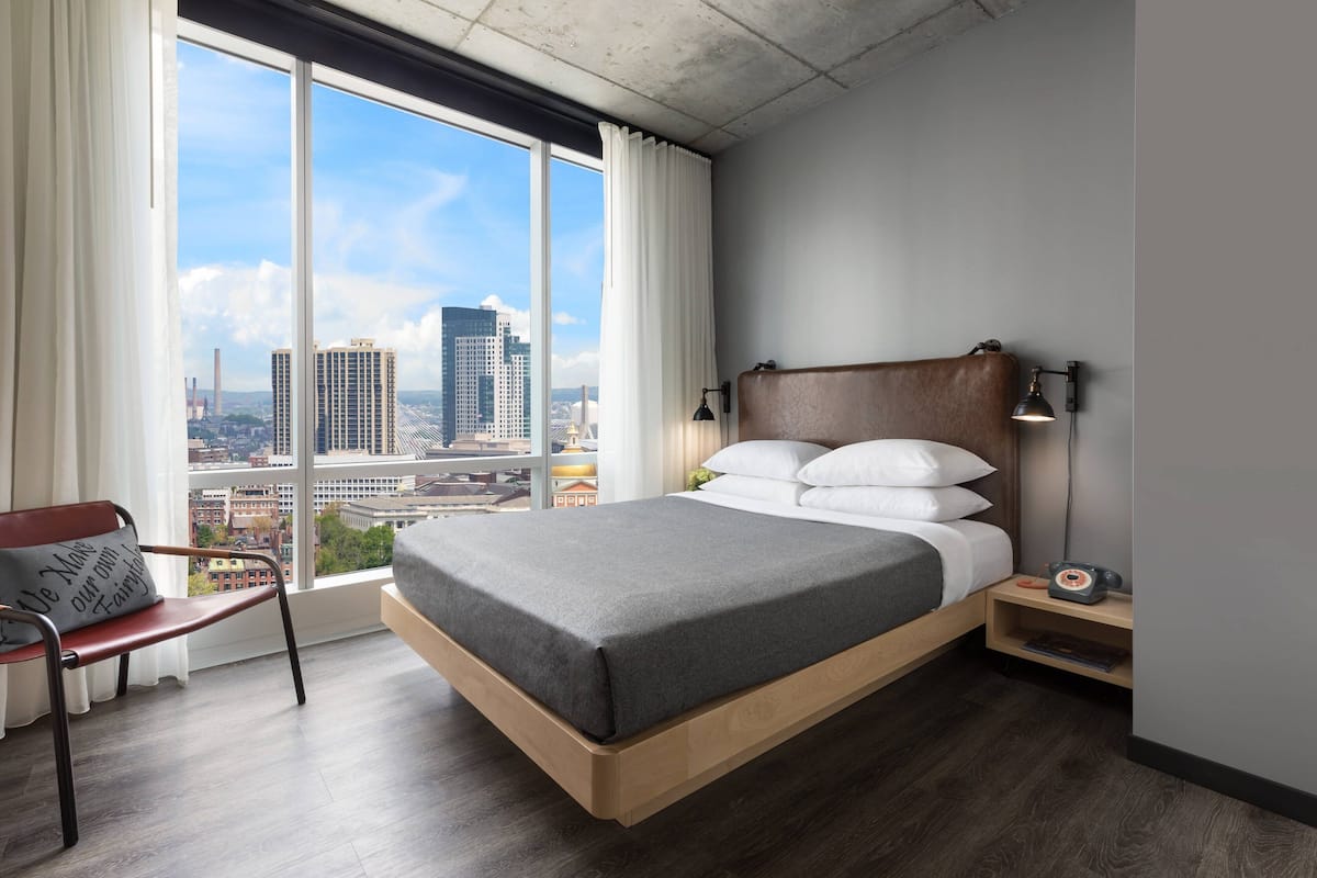 A modern hotel room with a large window, a wooden bed, a nightstand, and a chair.