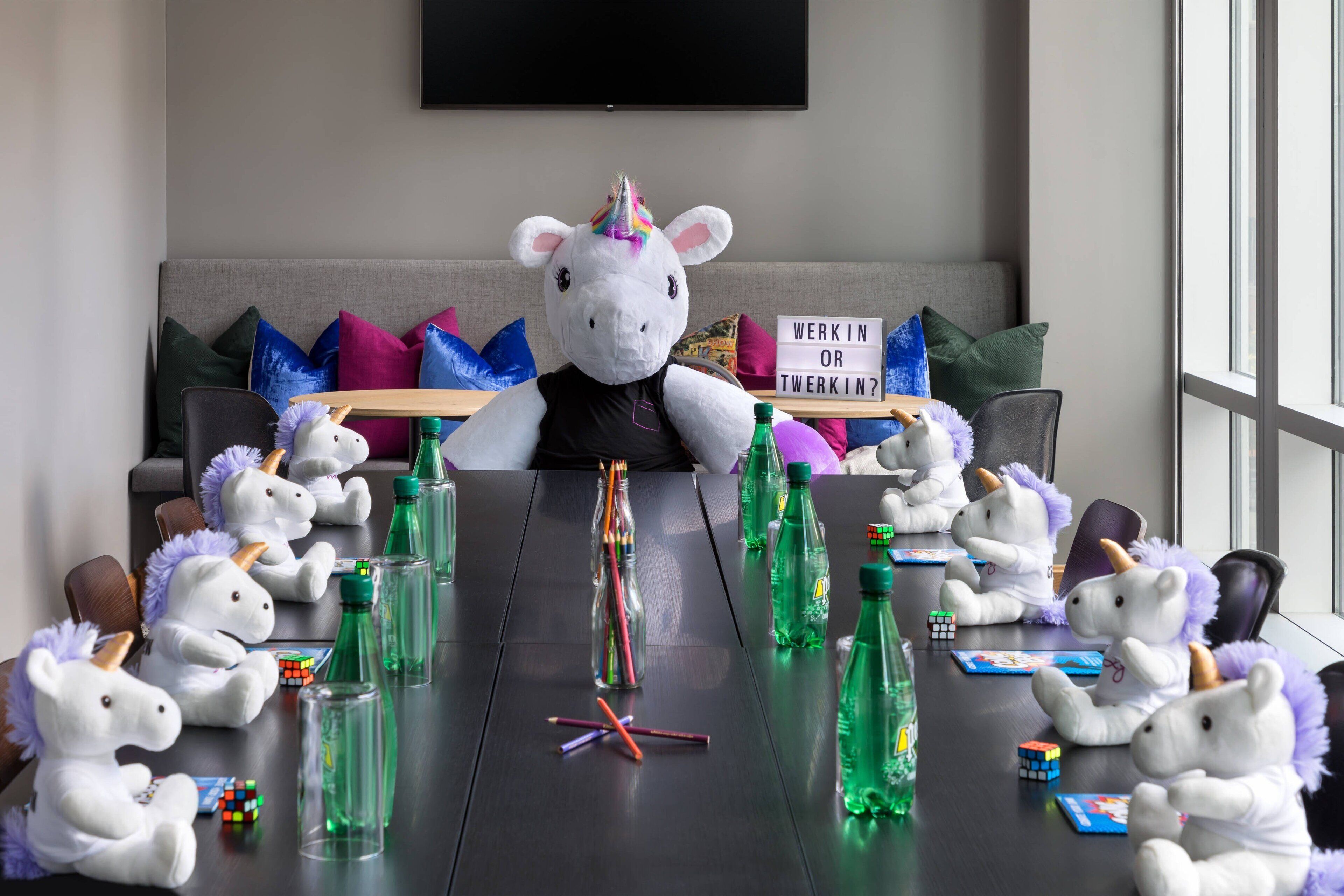 A meeting room with a table set for a meeting, featuring unicorn plush toys, a Rubik's Cube, and green bottles.