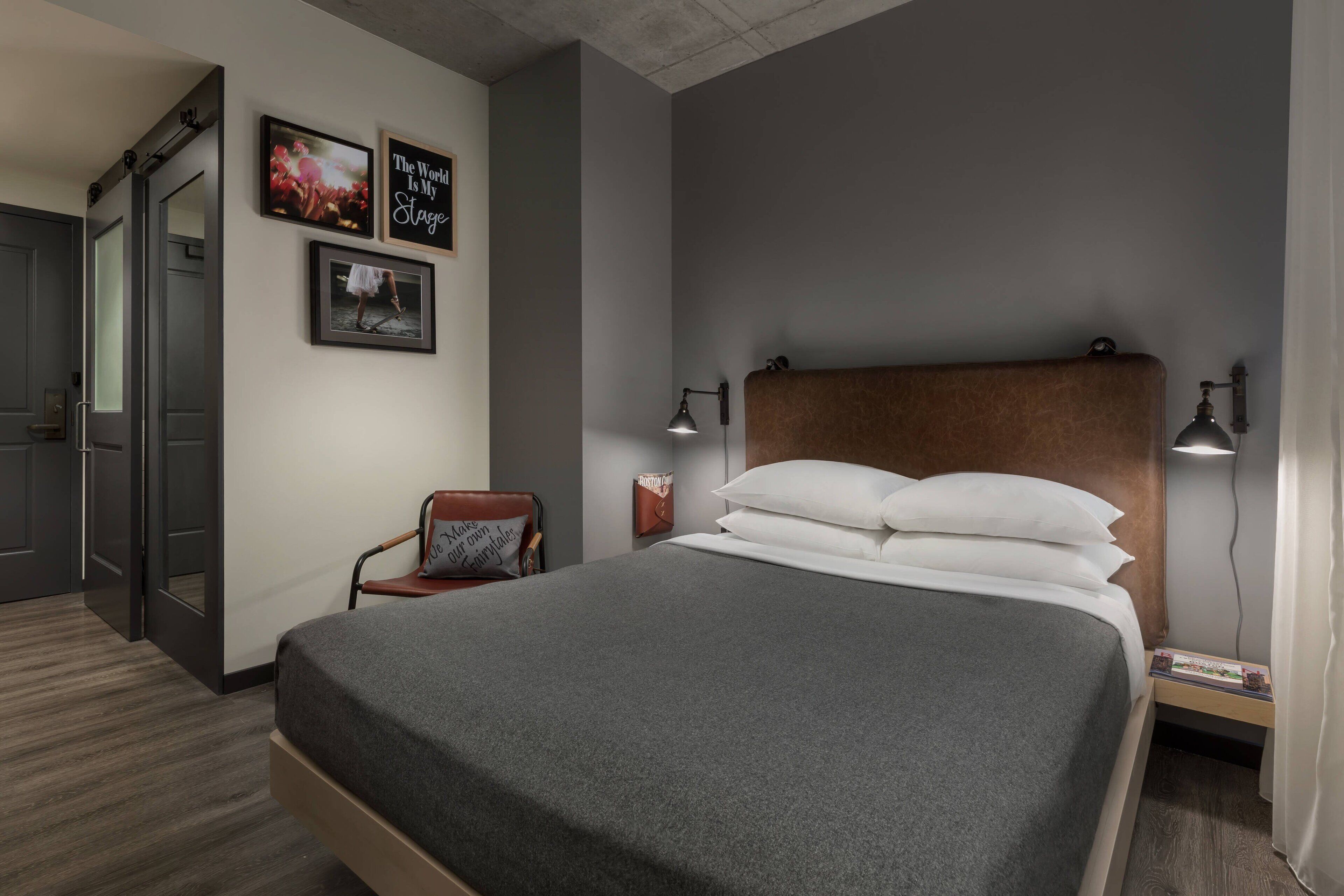Una habitación de hotel ordenada con una cama, una silla, una mesita de noche y una pared decorada con cuadros.