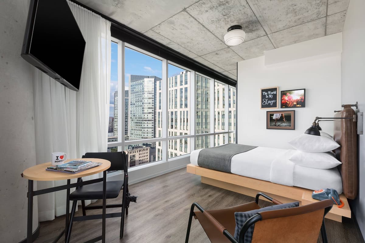 A modern hotel room with a bed, a desk, and a chair. There are large windows with a city view.