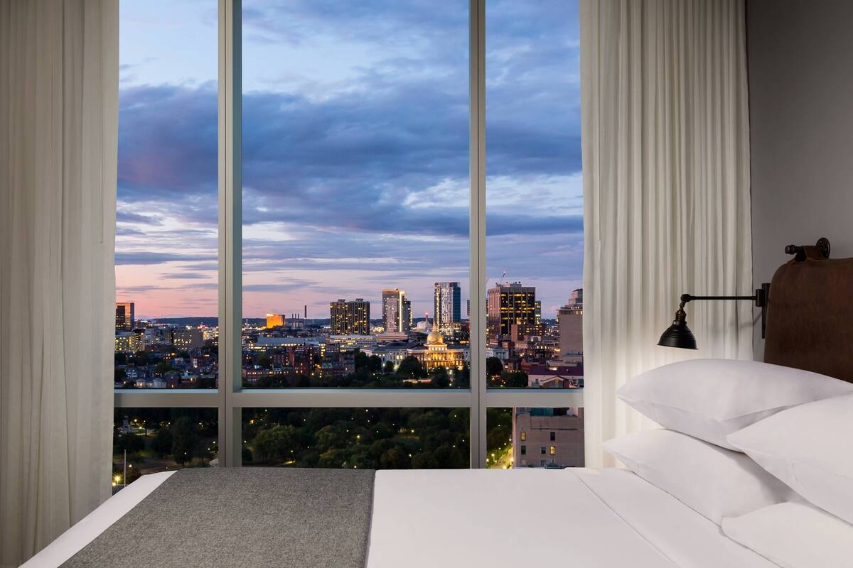 A hotel room with a large window offering a cityscape view, a bed, and a nightstand with a lamp.