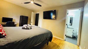 Un dormitorio con cama, escritorio, silla, televisión y baño visible a través de una puerta abierta.