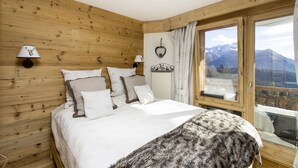 A bedroom with a wooden wall, a bed with white bedding, two wall-mounted lamps, a heart-shaped decoration, and a window with a mountain view. - Villars Bristol Apartments (Ollon)