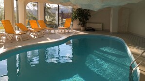 An indoor swimming pool with clear blue water, surrounded by white lounge chairs and large windows offering a view of the outdoors. - Villars Bristol Apartments (Ollon)