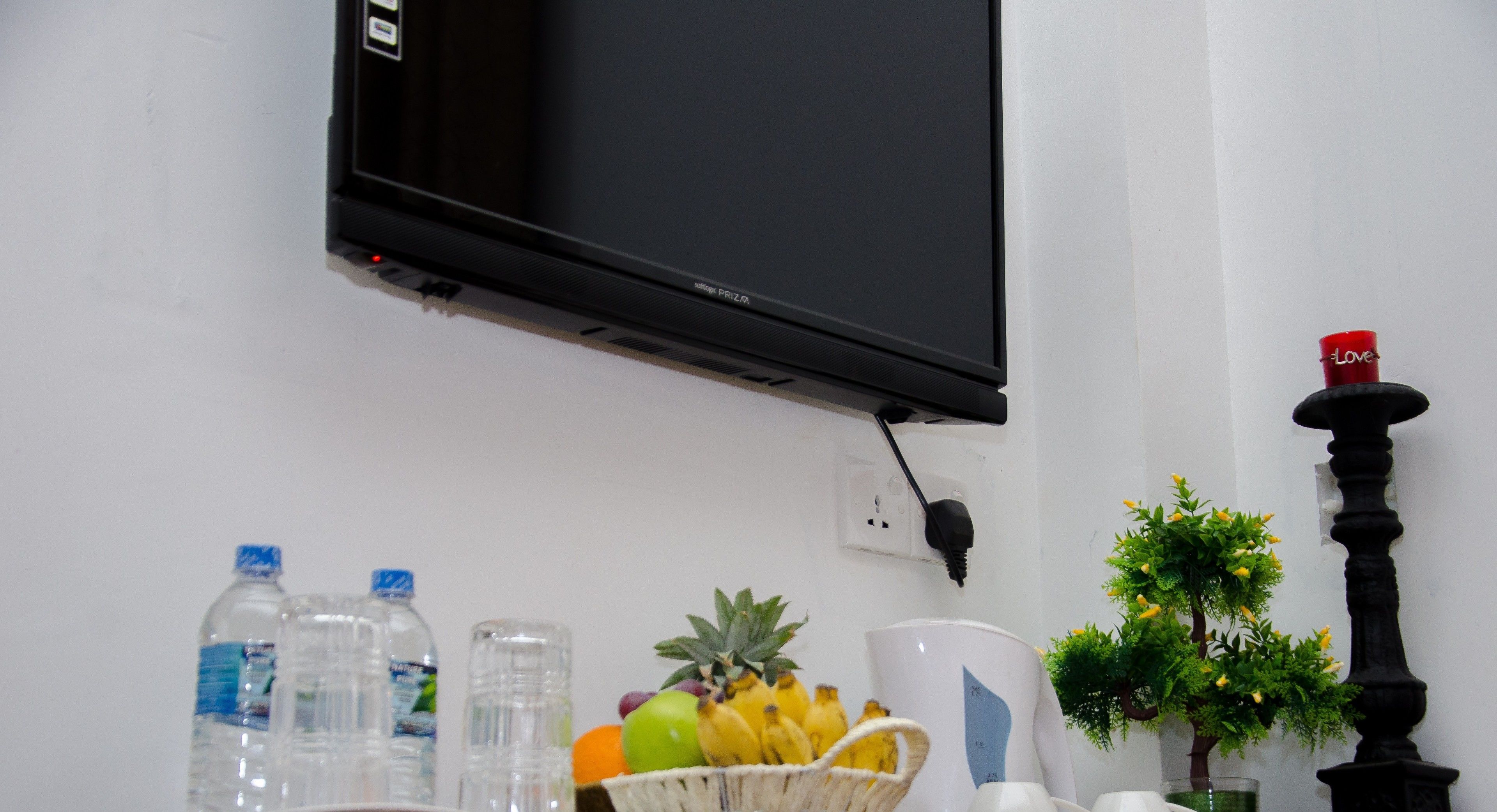 Un téléviseur à écran plat fixé au mur, une petite plante en pot, un pilier noir décoratif et un panier contenant des fruits et une tasse.