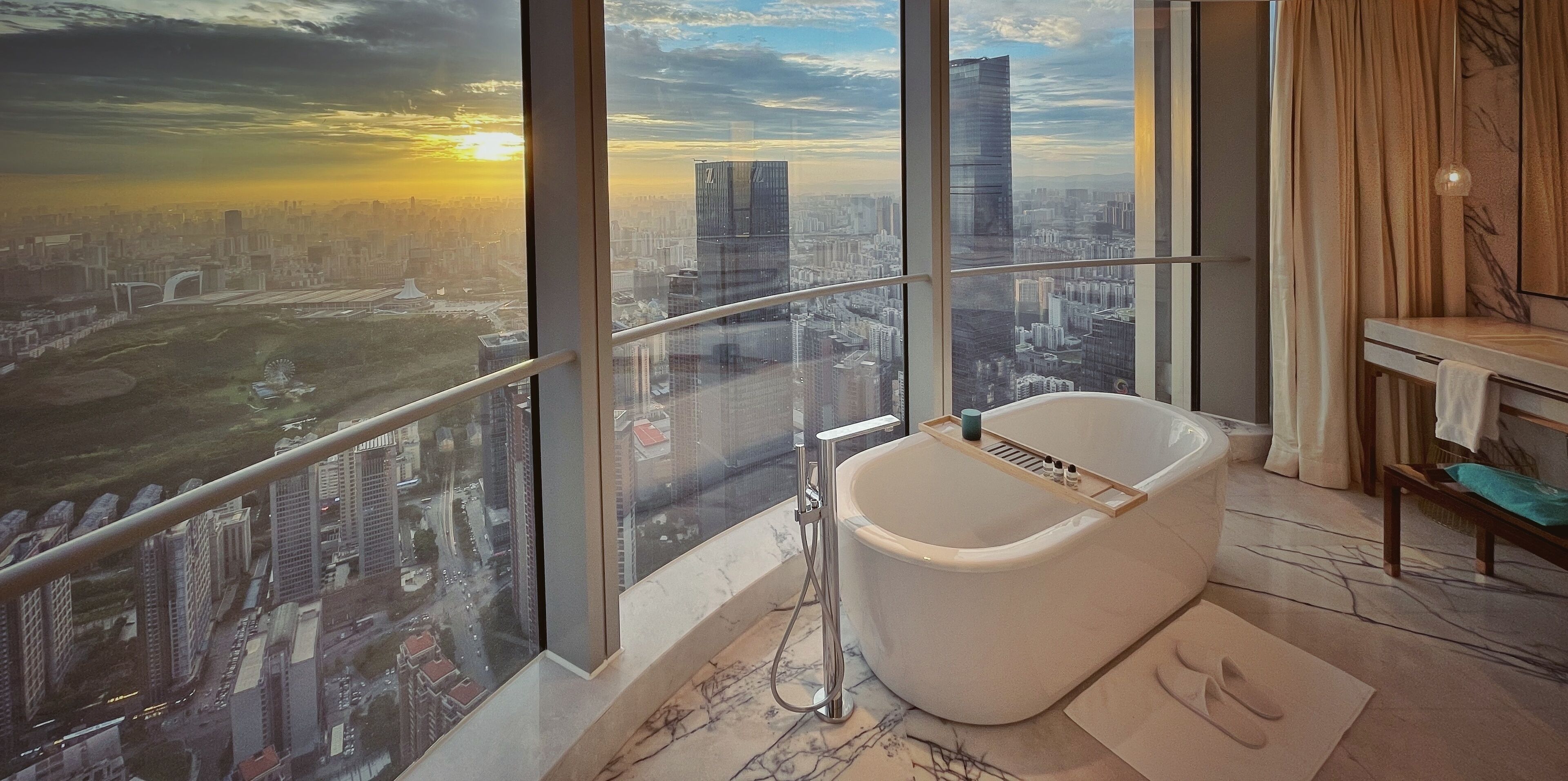 Un baño moderno con una bañera grande, vistas de un paisaje urbano a través de amplias ventanas y una vista de la ciudad mediante una barandilla de vidrio.