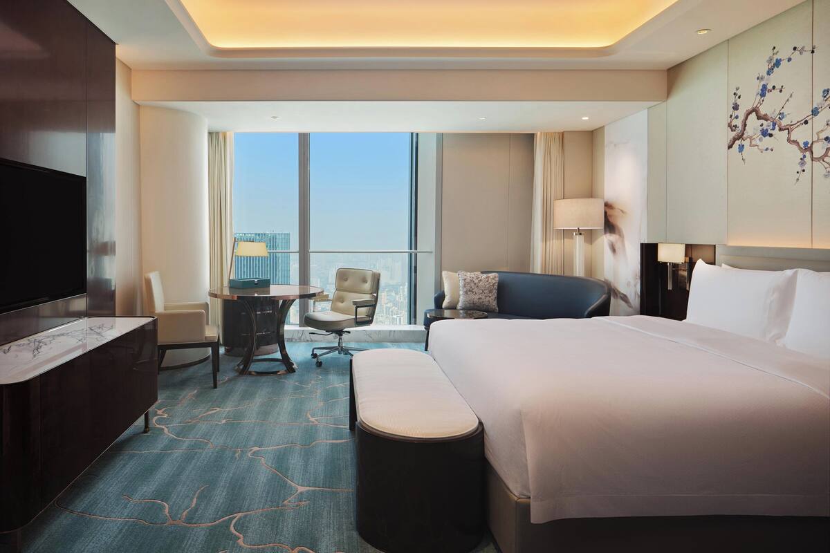 A hotel room with a large bed, a desk, a chair, a TV, and a view of the city through large windows.