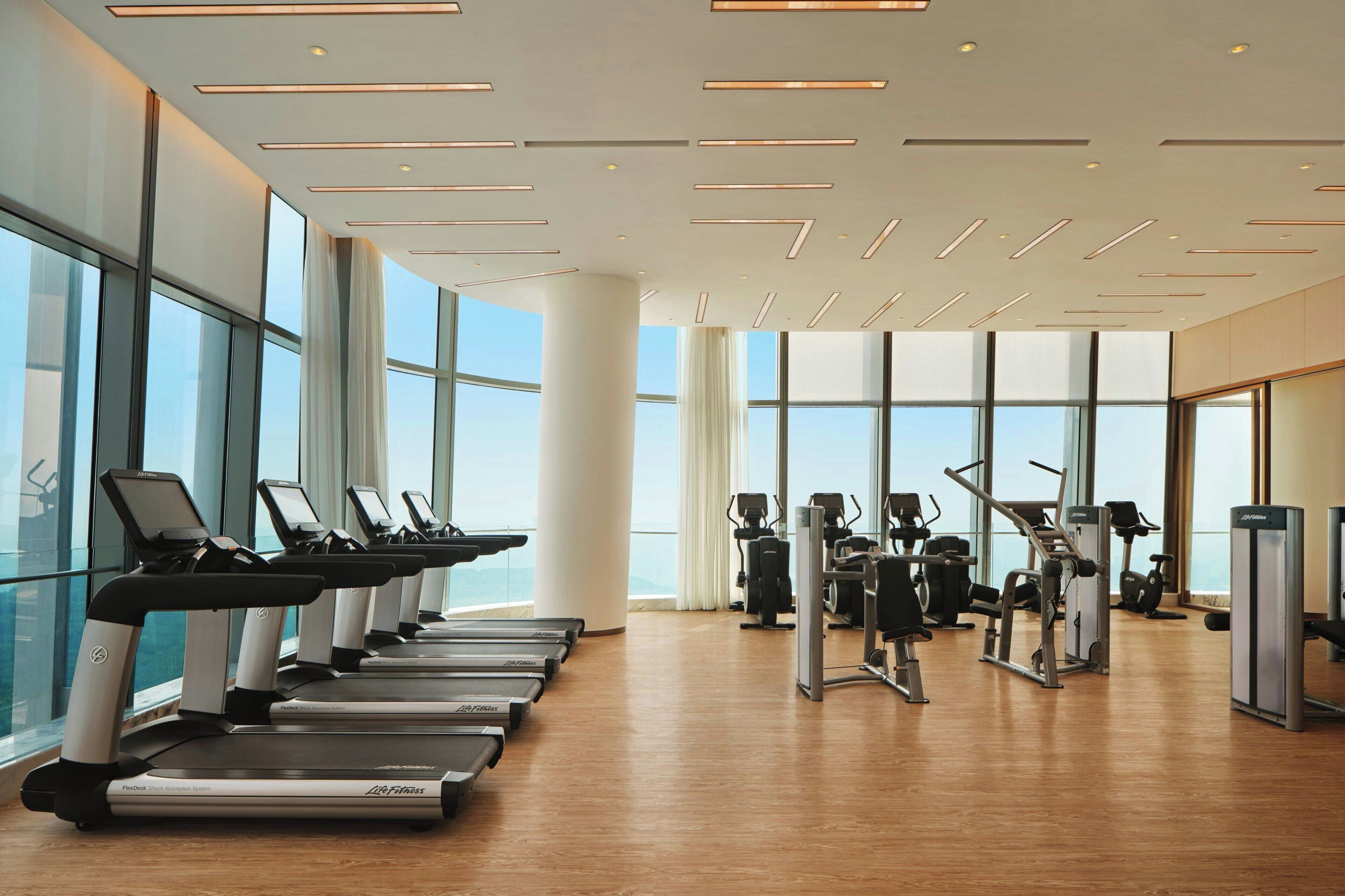 Un gimnasio con cintas de correr, máquinas de ejercicio y amplios ventanales que ofrecen vistas de la ciudad.