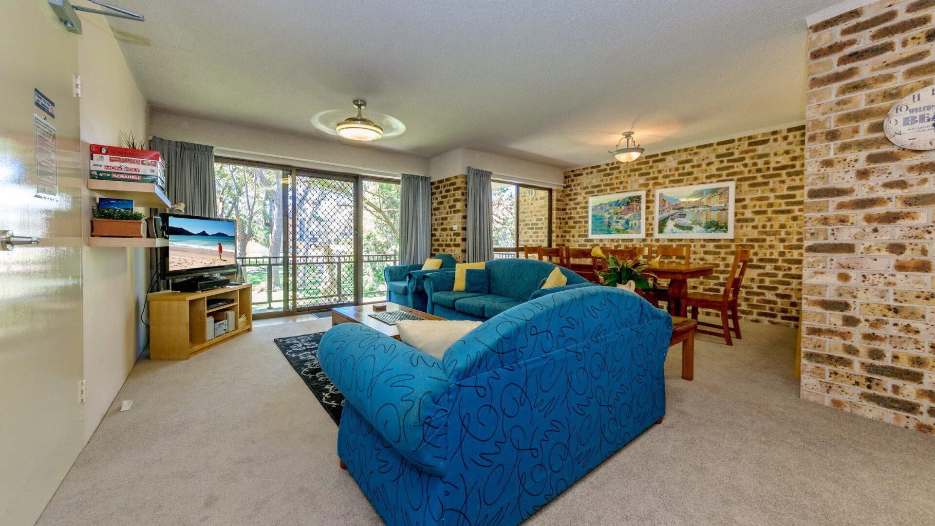 A living room with a blue sofa, a TV, and a dining area with a brick wall and two paintings.