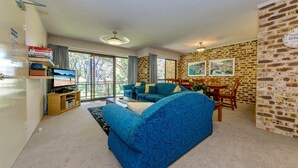 A living room with a blue sofa, a TV, and a dining area with a brick wall and two paintings. - Bay Parklands, 28/2 Gowrie Ave - pool, tennis + stunning views (Nelson Bay)