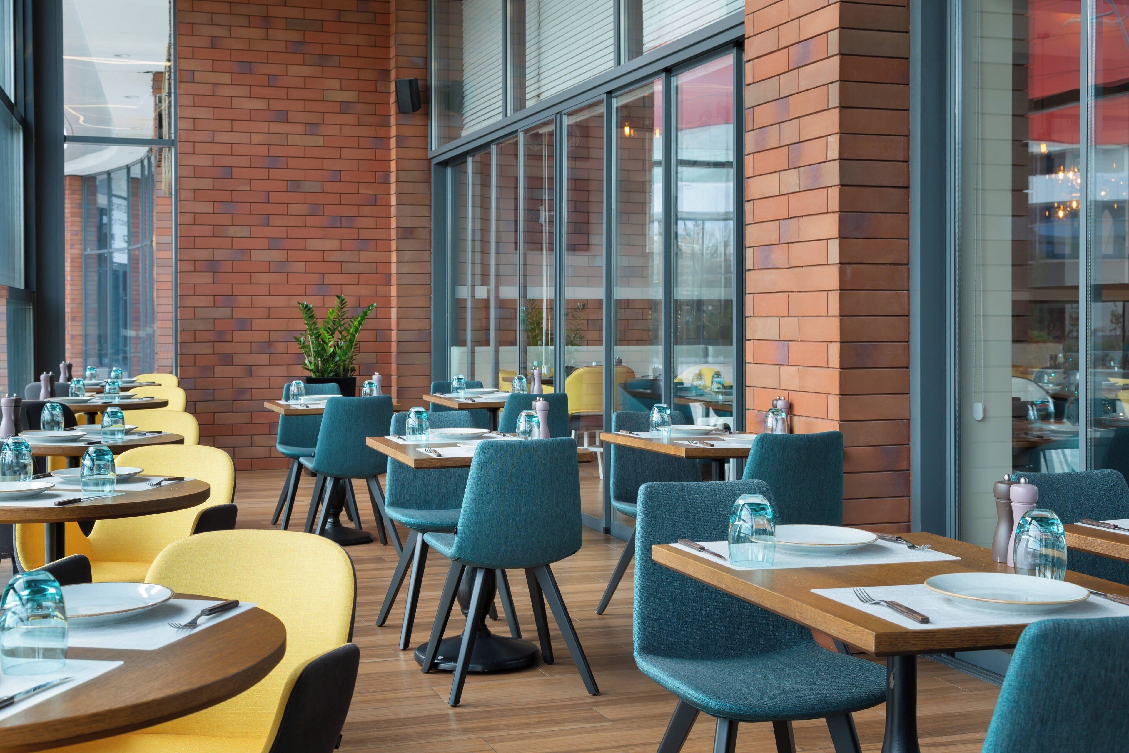 Ein Restaurant mit mehreren Tischen und Stühlen, großen Fenstern und einer Backsteinwand.