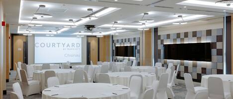 Un salón de conferencias con varias mesas redondas y sillas blancas, una pantalla grande mostrando 'COURTYARD by Marriott Chisinau', y un diseño de techo moderno.