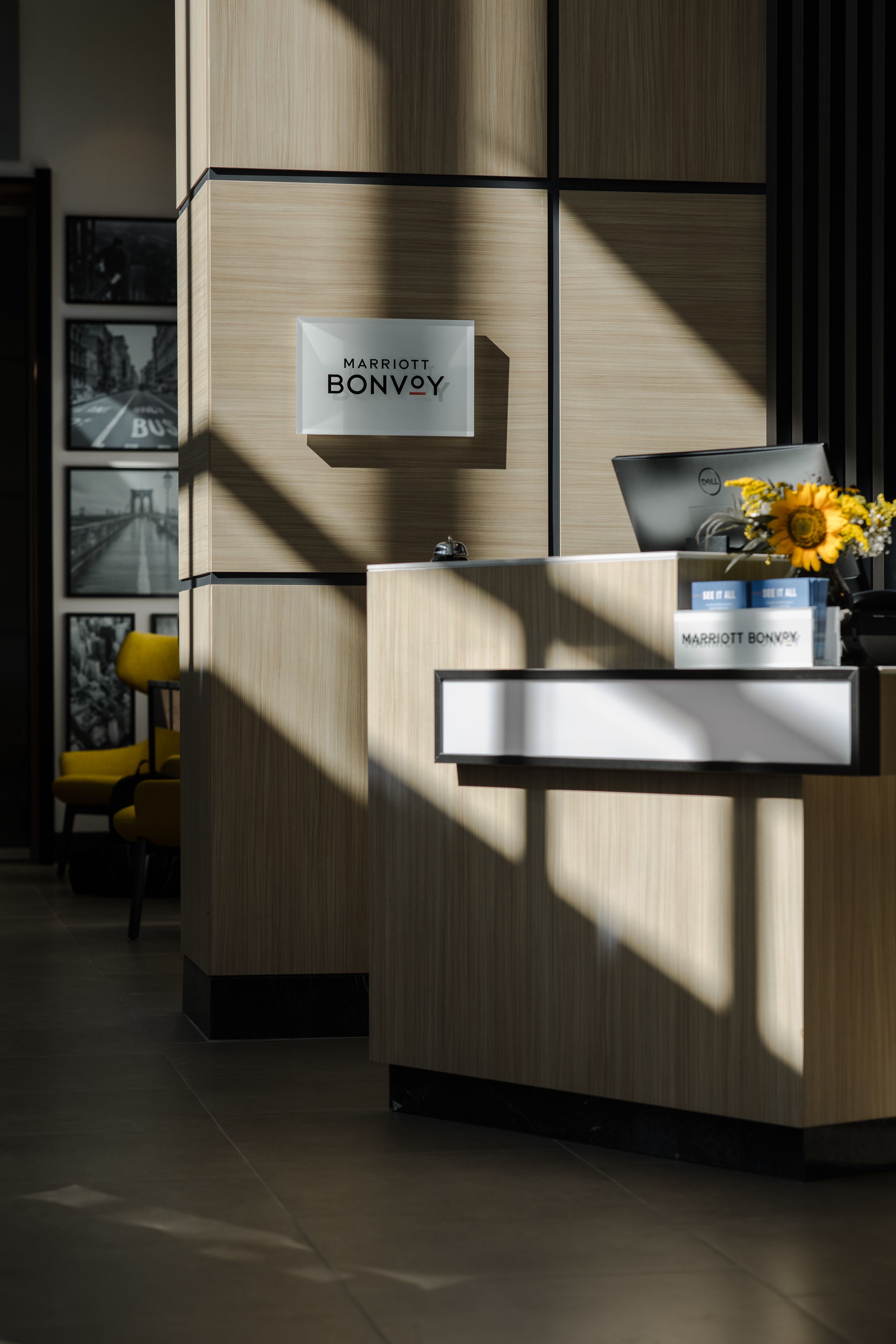 A reception desk at a Marriott Bonvoy hotel with a laptop, a vase with sunflowers, and framed pictures on the wall.