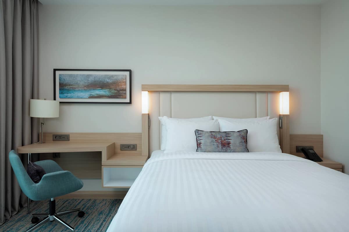 Quarto de hotel com cama, escrivaninha, cadeira, pintura e mesa de cabeceira com telefone.