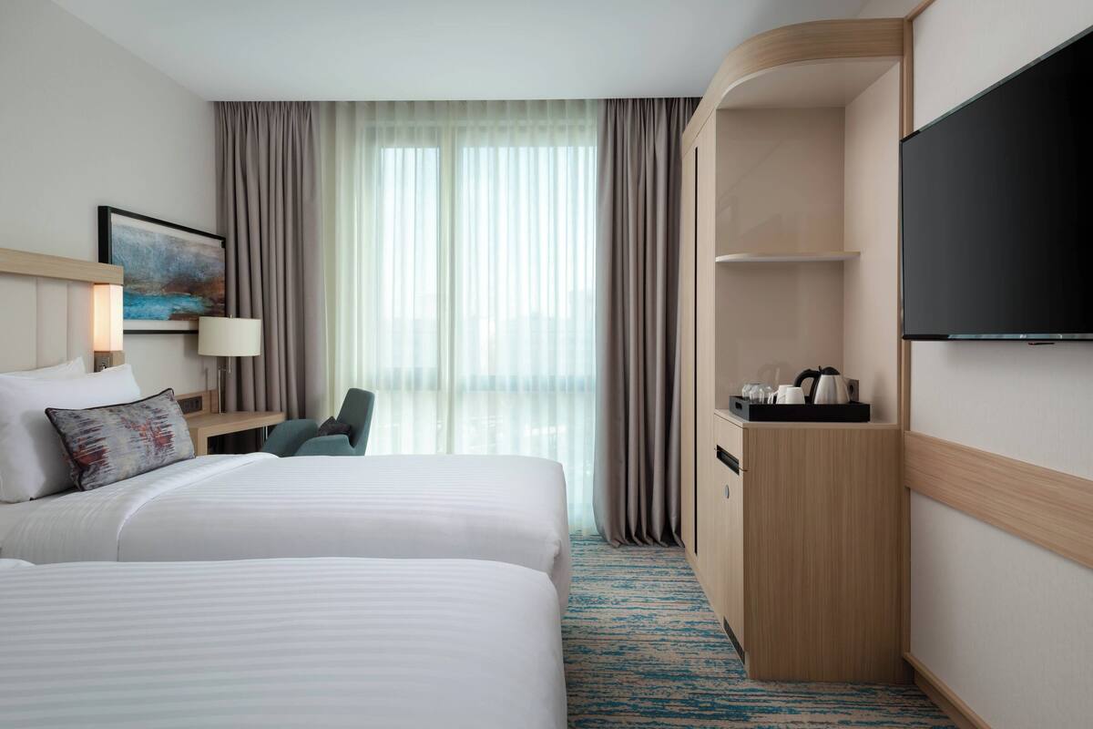 Quarto de hotel com duas camas, uma escrivaninha, uma cadeira, uma televisão e uma janela com cortinas.