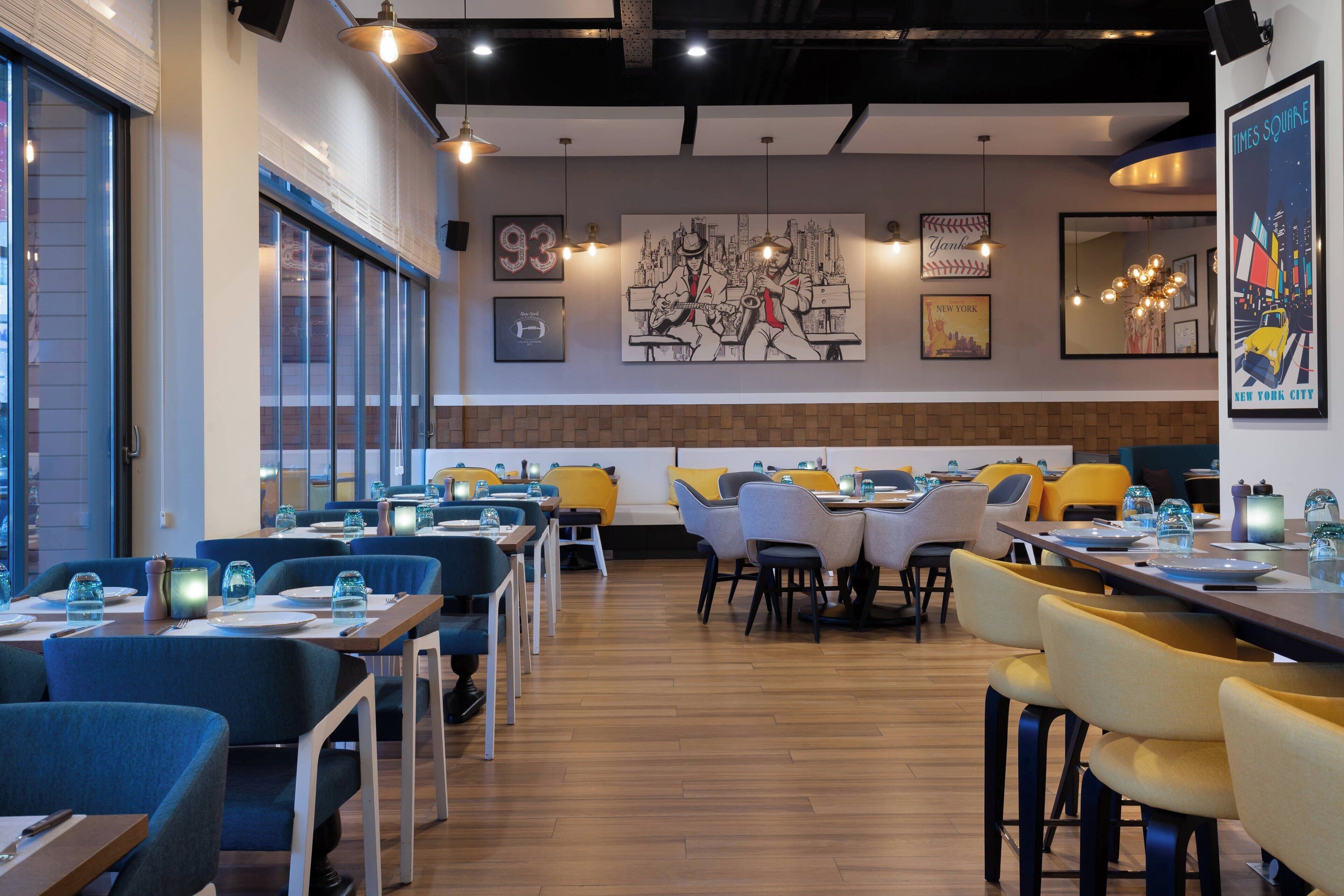 Interior moderno de restaurante con mesas y sillas, ventanas amplias y obras de arte en las paredes.
