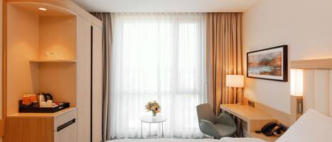 A hotel room with a bed, a desk with a lamp, a chair, a small table with a vase, and a painting on the wall.