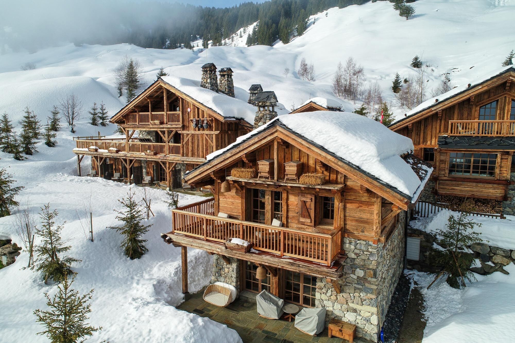 A wooden chalet with a balcony and stone chimneys, surrounded by snow-covered trees and a mountainous backdrop.