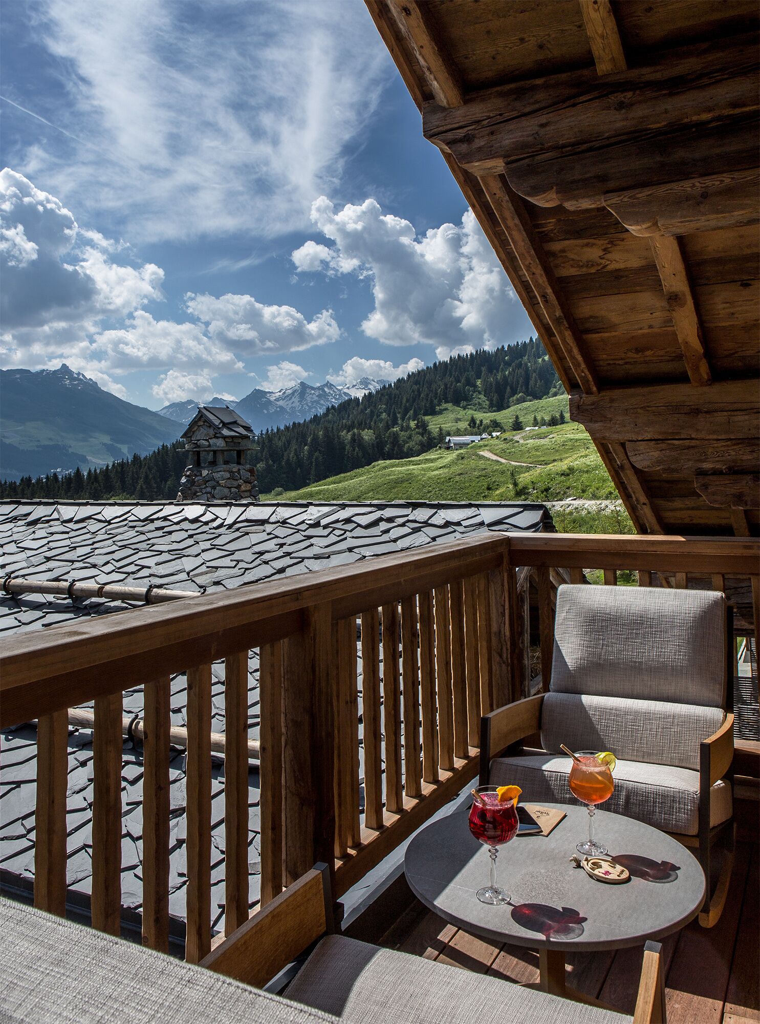 A wooden deck with a table and chairs, overlooking a mountainous landscape with a stone building.