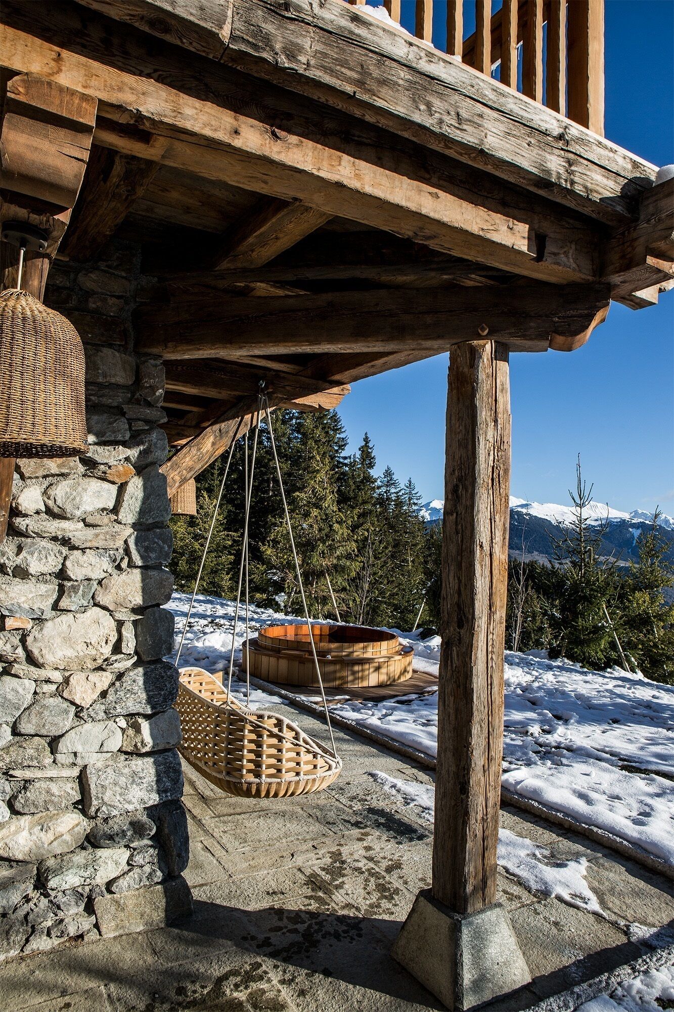 Un balcon en bois avec des murs en pierre, une jardinière suspendue et une cuve en bois, le tout posé sur un sol enneigé, avec des arbres et des montagnes en arrière-plan.