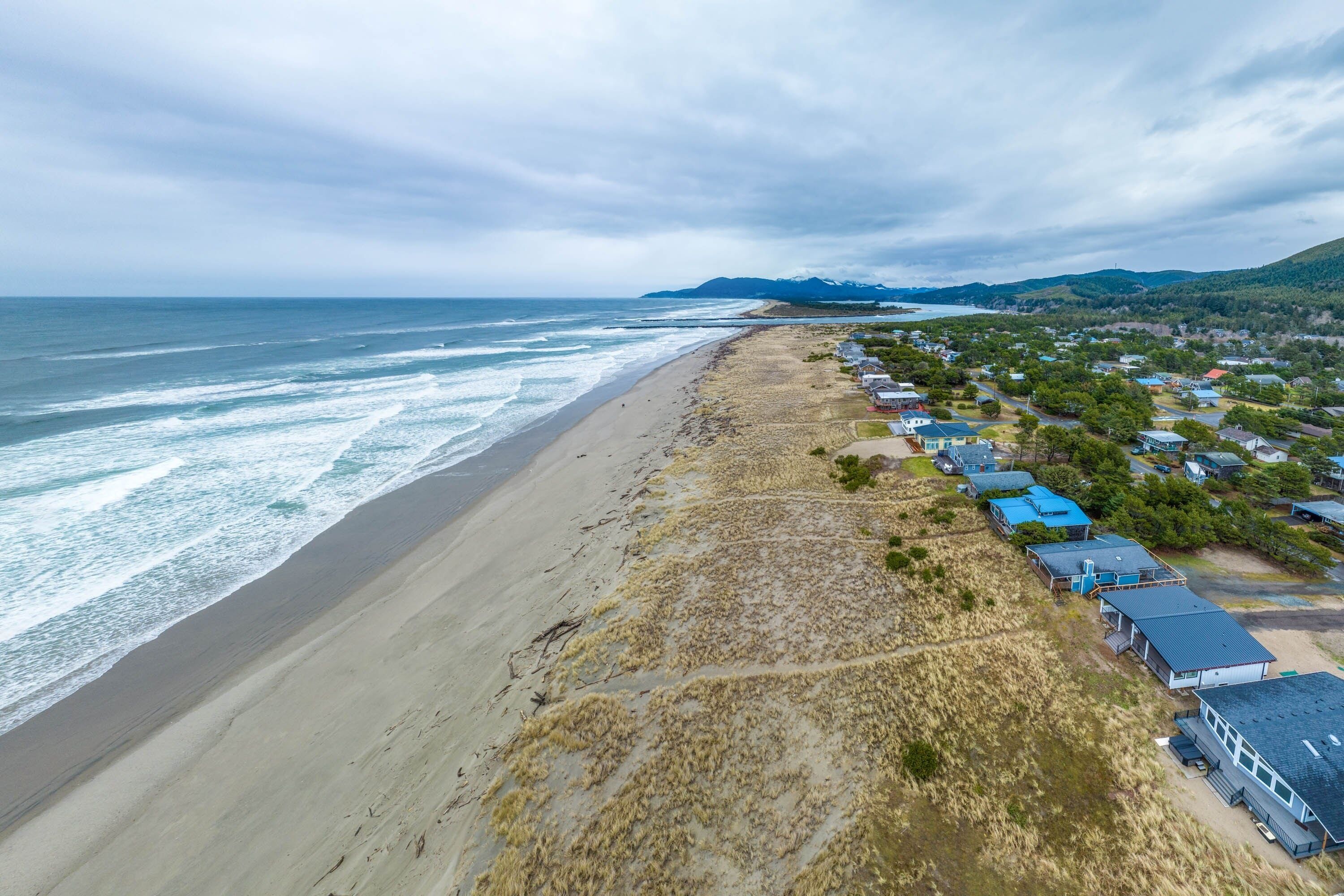 Una zona costera con una playa arenosa, una hilera de casas con tejados azules y un paisaje montañoso al fondo.