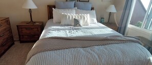 Un lit bien fait, recouvert d’une couverture matelassée, deux tables de chevet avec des lampes, une commode en bois et une fenêtre avec des rideaux.