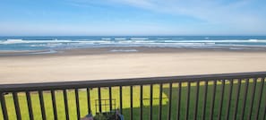 A beach view from a balcony with a railing, a grassy area, and a clear sky.