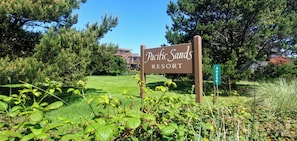 A sign for Pacific Sands Resort surrounded by greenery and a building in the background.