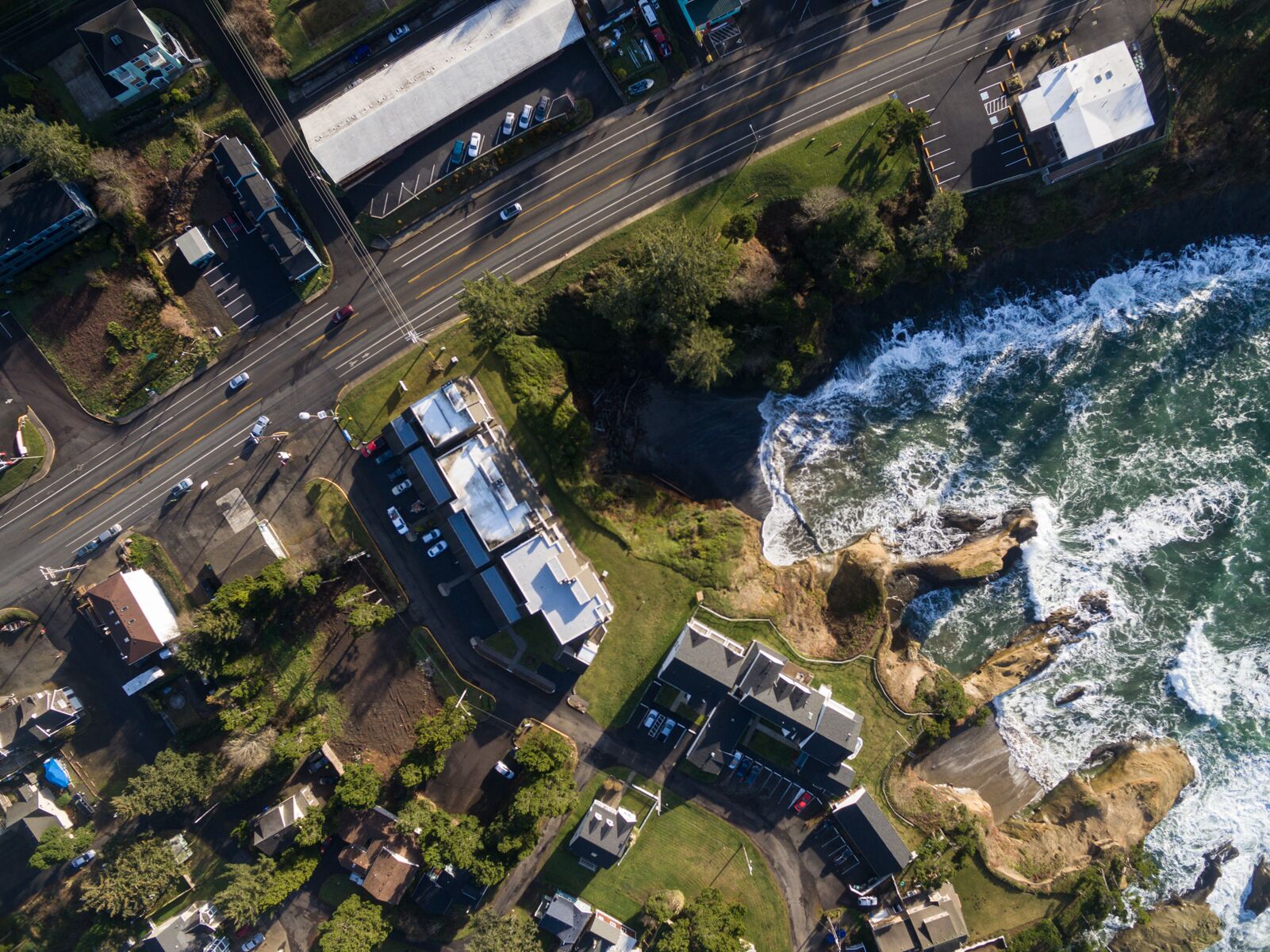 An aerial view of a coastal area with buildings, roads, and a rocky shoreline with waves.