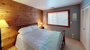 A bedroom with a wooden headboard, a bed with patterned bedding, a bedside lamp, a window with blinds, and a door with shutters.