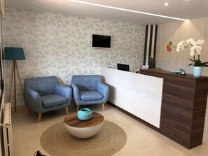 A reception area with a wooden reception desk, two blue armchairs, a round coffee table, a flat-screen TV mounted on the wall, and a potted orchid on the desk. - Hotel Florida (Sanxenxo)