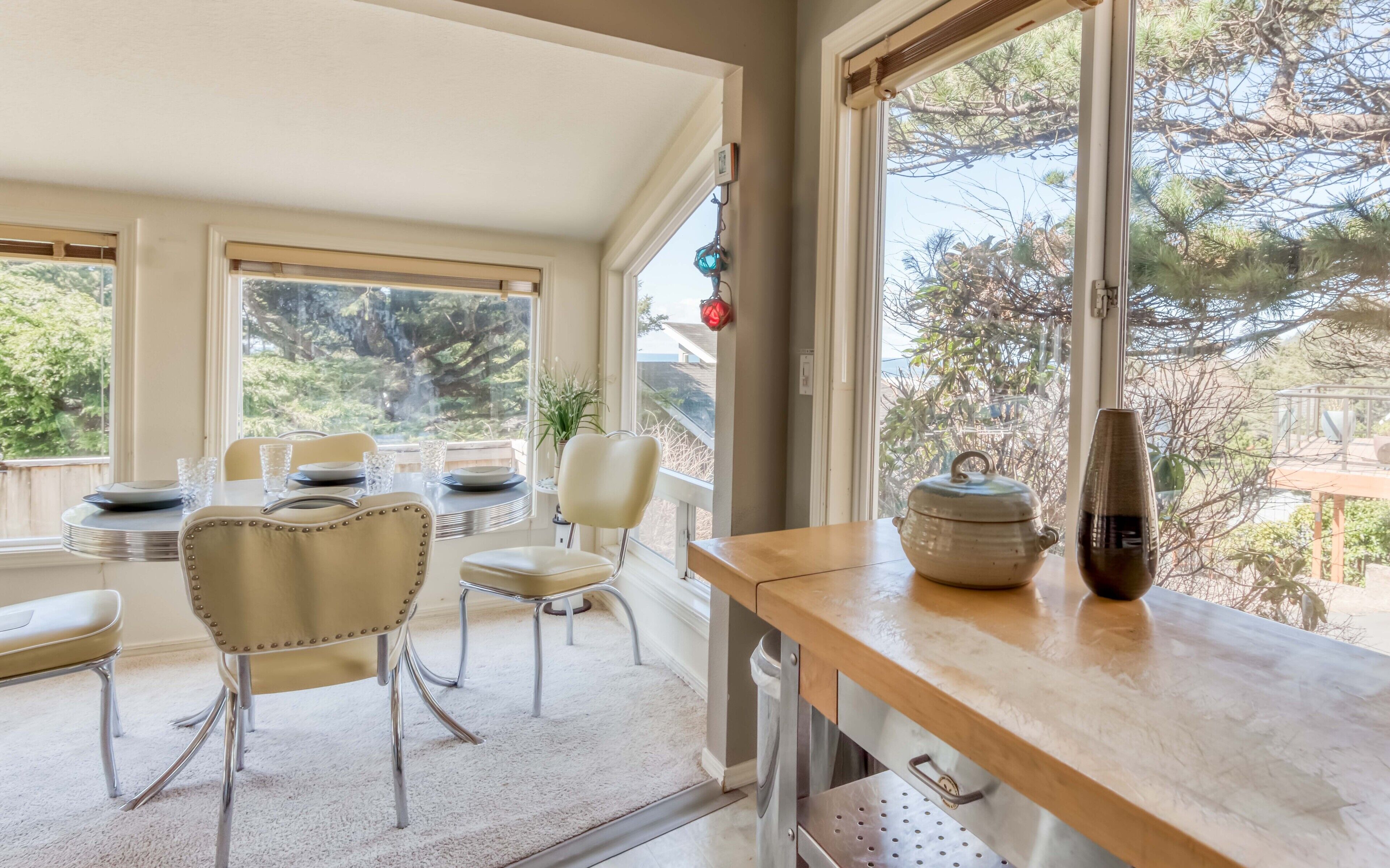 A dining area with a table set for four, chairs, a vase, and a window with a view of trees and a building.