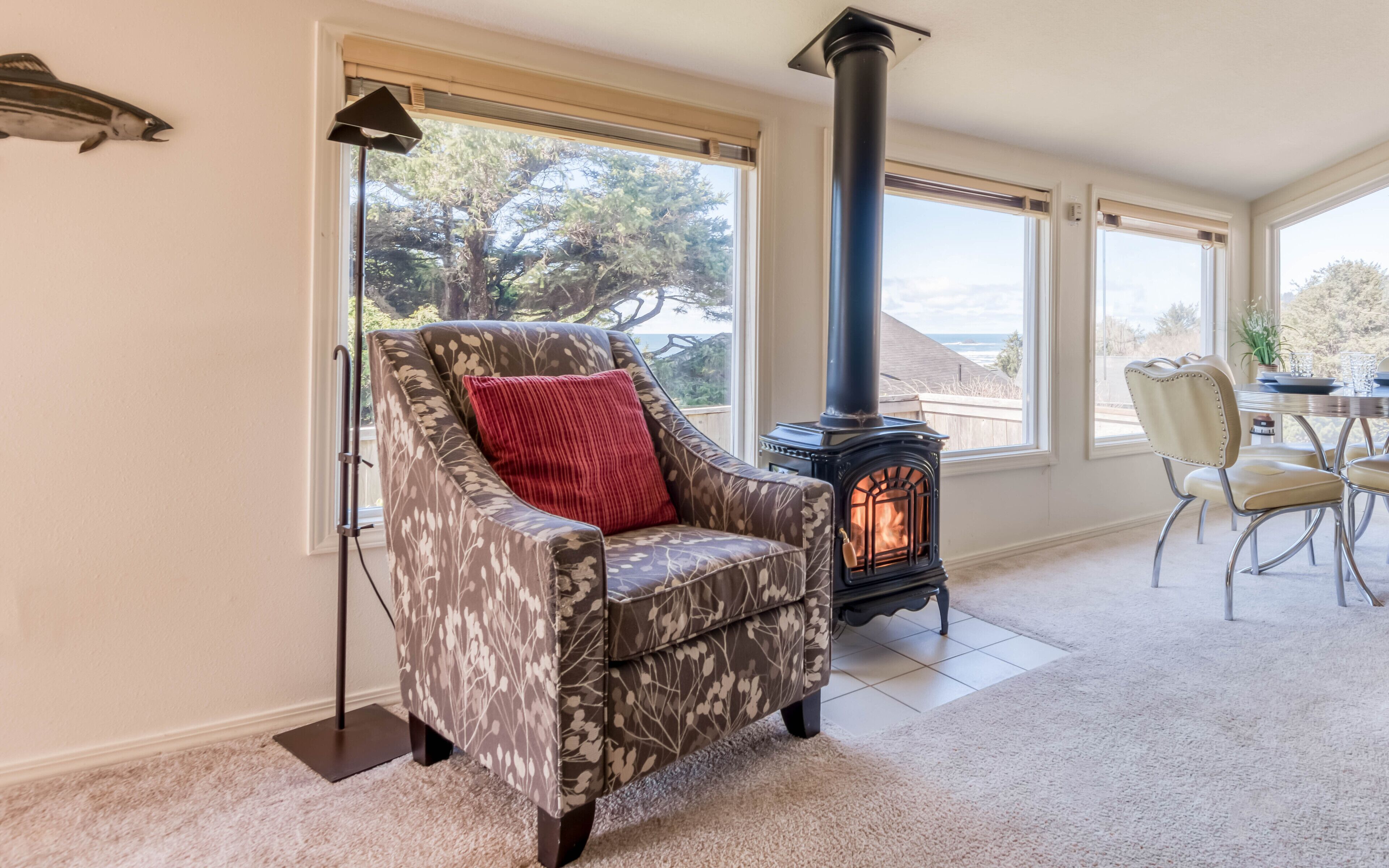 A cozy living room with a patterned armchair, a red pillow, a black wood-burning stove, and large windows offering a view of the outdoors.