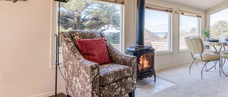 A cozy living room with a patterned armchair, a red pillow, a black wood-burning stove, and large windows offering a view of the outdoors.