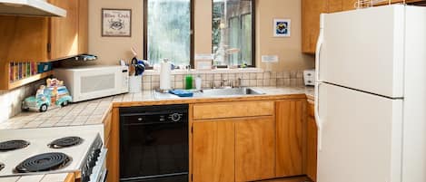 Una cocina con armarios de madera, un refrigerador blanco, una estufa y un lavavajillas. Hay dos ventanas con vistas al exterior, con abundante vegetación.