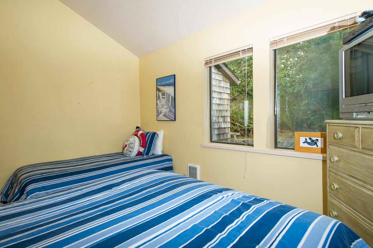 A bedroom with two beds, a dresser, a television, and a window with a view of greenery.