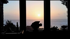 Une vue sur l’océan au coucher du soleil, prise à travers une fenêtre, avec la silhouette d’un oiseau et d’un phare en arrière-plan.
