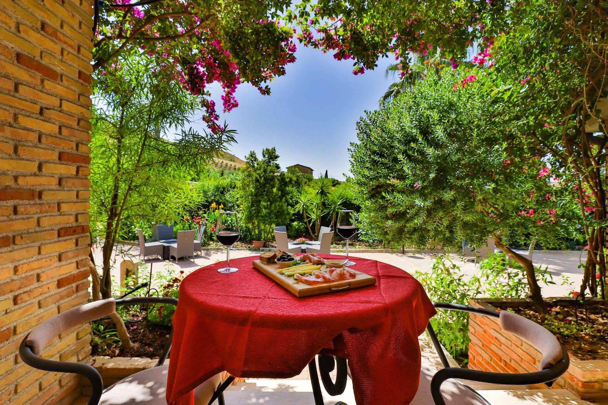 Un patio exterior con una mesa lista para comer, rodeado de follaje y un cielo despejado.