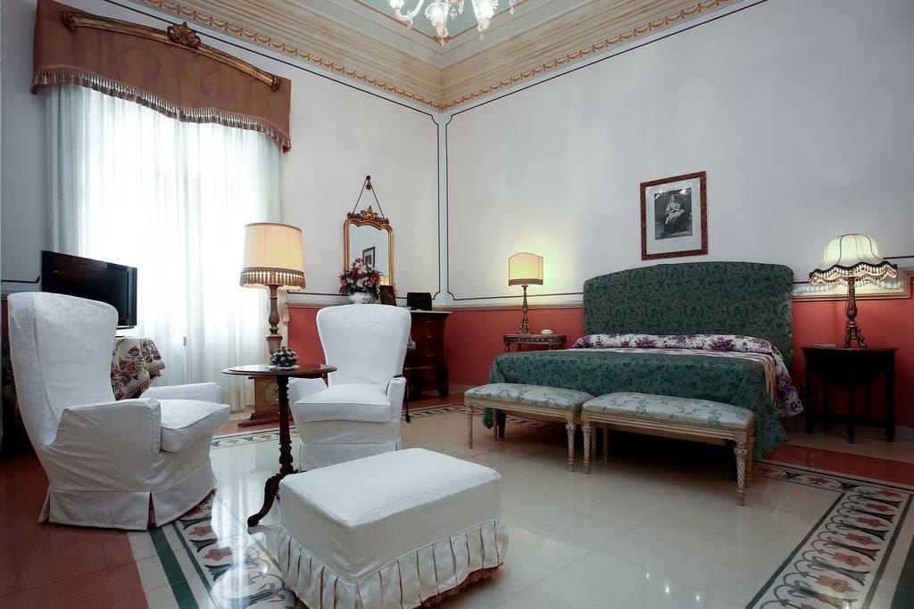 A hotel room with a large bed, two white armchairs, a small table, a bench, a TV, and a chandelier.