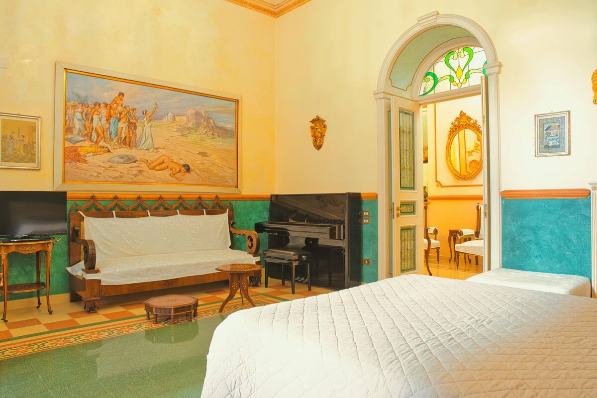 A hotel room with a large bed, a sofa, a piano, a painting, and a mirror.